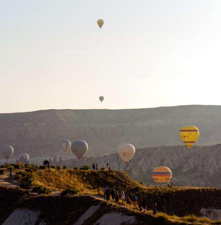 Goreme, Cappadocia, Turkey â 22 August 2019: Many hot air balloons fly in sky at dawn. People look at them from the ground. Cappadocia Earth Pyramids. Tourism concept.のeditorial素材