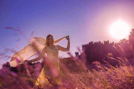 Blonde woman in a yellow dress in lavender field at sunset. Lavender bloom season. Enjoyment of unity with nature. Meditation and relaxation outdoorの写真素材