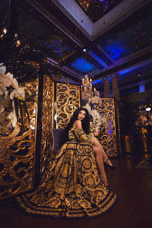 Portrait of a beautiful tanned brunette girl in a silk black and gold dress in a luxurious interior with gold decor, crystal and orchidsの写真素材