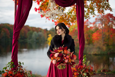 A beautiful bride in a dark red dress stands under a large single tree with red and yellow leaves. A destination autumn wedding, romantic country ceremony with marsala decor.の写真素材