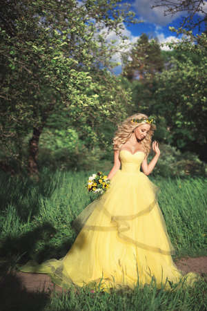 A gorgeous blonde girl in a lush yellow ball gown with a pansies bouquet and wreath walking in a summer garden among green trees and grasses. Romantic sunny portrait.の写真素材