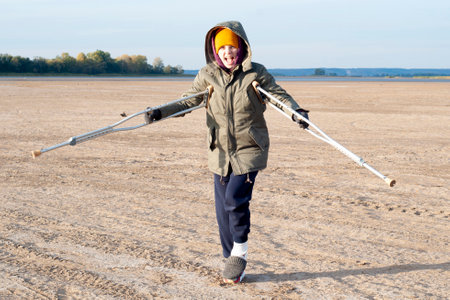 A young girl broke her leg. It stands in nature, on the street, on crutches on vacation. The concept of rest with a broken leg. In cold autumn, a young woman walks on crutches in nature.の写真素材