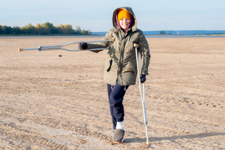 A young girl broke her leg. It stands in nature, on the street, on crutches on vacation. The concept of rest with a broken leg. In cold autumn, a young woman walks on crutches in nature.の写真素材