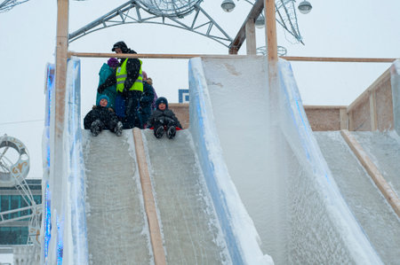 Small children slide down a snow-covered slope, in the winter on the street, ride down a winter slide. Skating on slippery surfaces outdoors. folk fun. Street New Year holidays. city hill.のeditorial素材