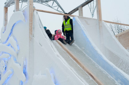 Small children slide down a snow-covered slope, in the winter on the street, ride down a winter slide. Skating on slippery surfaces outdoors. folk fun. Street New Year holidays. safety equipment.のeditorial素材