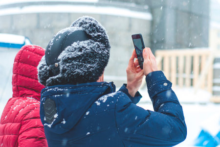 A man in a winter jacket, in a winter fur hat, without a face, stands with his back and holds a phone, a smartphone in his hands, in the cold, in the cold, in winter. winter day.の写真素材