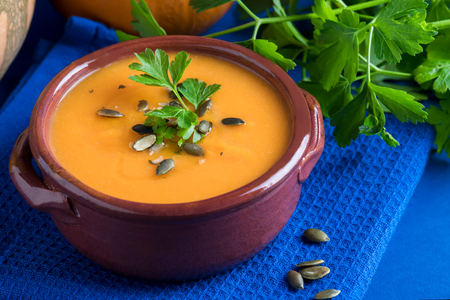 Close up of pumpkin vegan soup in a clay bowl served with parsley, olive oil and pumpkin seeds on the blue backgroundの写真素材