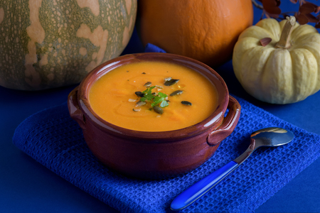 Pumpkin vegan soup in a clay bowl served with parsley, olive oil and pumpkin seeds on the blue backgroundの写真素材