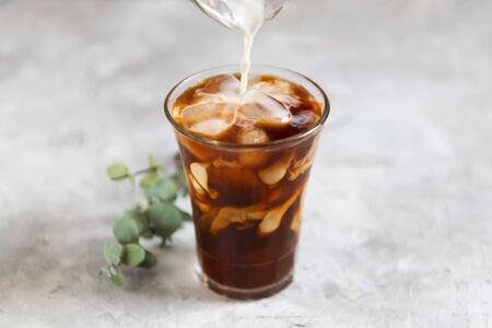 Milk is pouring into a tall glass with brew black coffee with pieces of ice on a gray dark background, close-up. Ice summer fresh coffee conceptの写真素材
