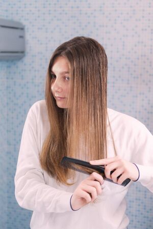A beautiful girl is combing her hair herself, looking in mirror in the bathroom, morning routine,...