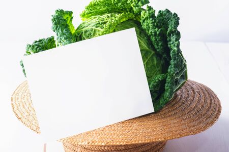 Grenery mock up invitation card or baby card with fresh savoy cabbage in a straw hat on white wooden background, creative food, copy spaceの写真素材