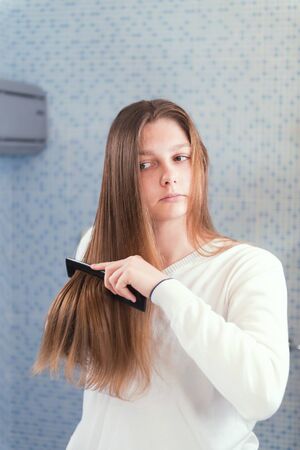 A beautiful blond girl is combing her hair herself, looking in mirror in the bathroom, morning routine, selective focusの写真素材