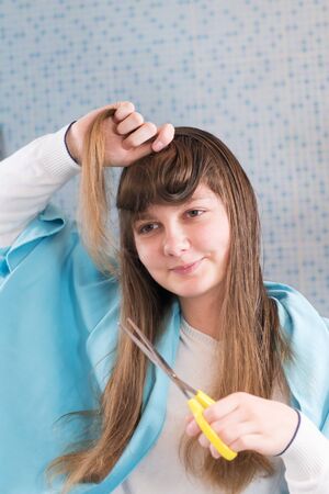 A beautiful young girl is holding a scissor to cut her hair herself during lockdown of coronavirus, looking in mirror. DIY hair cut during the epidemic of covid-19, stay at home, selective focusの写真素材