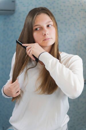 A beautiful girl is combing her hair herself, looking in mirror in the bathroom, morning routine, selective focusの写真素材
