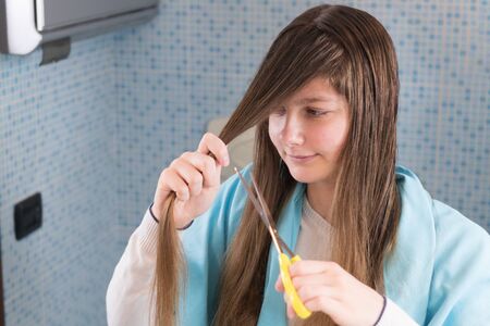 A girl is holding a scissor to cut her hair herself during lockdown of coronavirus, looking in mirror making a shocked and funny face. DIY hair cut during covid-19, stay at home, selective focusの写真素材