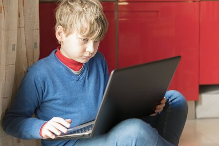A blond child boy is sitting on the floor in the room with laptop. Homeschooling and education from home online, space for textの写真素材