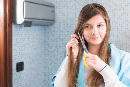 A beautiful young girl is holding a scissor to cut her hair herself during lockdown of coronavirus,...