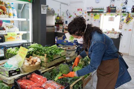 Avetrana, Italy, - Marth 16, 2020. An Italian greengrocer is taking broccoli and cabbage, wearing medical mask and gloves protection during Coronavirus epidemy. Shopping, pandemia of Covid-19のeditorial素材