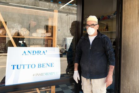 Avetrana, Italy, - Marth, 13, 2020. Italian baker with medical mask near an Andra tutto bene Everything will be ok in italian poster for the flashmob against Coronavirus. Shopping, pandemia Covid-19のeditorial素材