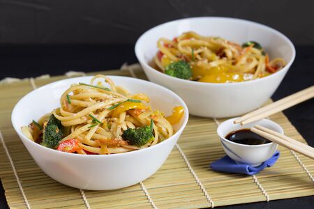 Asian food, two bowls of udon noodles with vegetables and soy sauce on a bamboo mat, black backgroundの写真素材