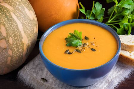 Pumpkin vegan soup in a blue bowl served with parsley, olive oil and pumpkin seeds and bread on a dark wooden backgroundの写真素材