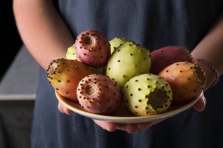 Female hands hold a plate with ripe fresh fruits of prickly pear on a gray background, low key, close up. Opuntia fruit grow in Puglia, Italyの写真素材