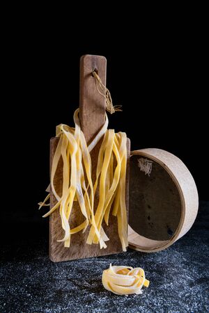 Italian homemade raw uncooked tagliatelle pasta hanging on a cutting board with a beautiful backlight on a black background, concept of Italian food, still life with a strainer and tagliatelleの写真素材