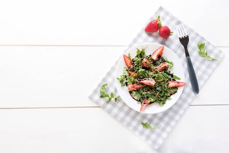 Healthy organic diet salad with arugula, strawberries and sesame in white plate on grey napkin on a white wooden background, scandinave style, healthy food clean eating concept, top view, copy spaceの写真素材
