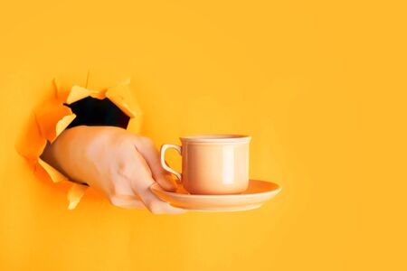 Hand holding a cup of coffee through a hole in torn saffron or light orange paper wall. Coffee break concept, copy space, minimalismの写真素材