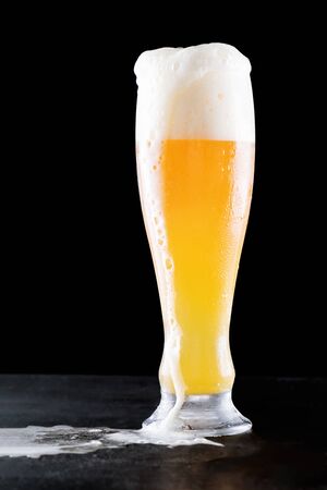 Cold Craft light lager beer in a overflowing glass with water drops with froth flowing and beautiful spilled beer on a black background with reflectionの写真素材
