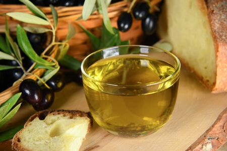 Healthy fats. Bowl of glass full of Extra Virgin Olive Oil made in Puglia, Salento on a wooden table with bread and olive branches. Tipical ingredients of mediterranean diet, healhty eating conceptの写真素材