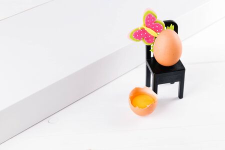 half raw egg with yolk on a white table and one egg is sitting on a black chair with easter flower...