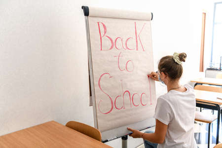 Young girl wearingmedical mask writing back to school on whiteboard in empty classroom, back to school during Covid-19 pandemic, new normal. Returning to school after quarantine, happiness and hope.の写真素材