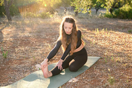 Young girl is doing yoga strethching on the mat on the olive grove at sunset. Female yoga training...