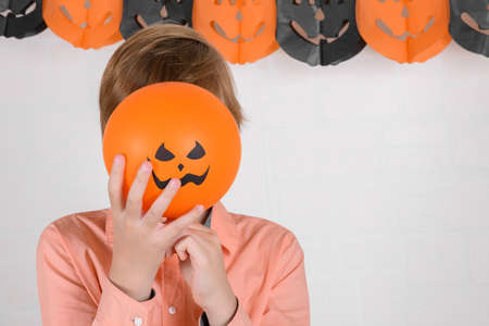A boy hides his face behind an orange halloween balloon. Celebrate Halloween on a white brick wall background. Happy Halloween on quarantine coronavirus pandemicの写真素材