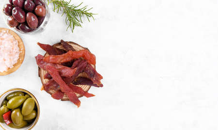 Jerky meat strips with spices, green olives, rosemary on wooden plate on a gray stone background....