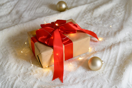 Kraft gift box with red ribbon bow on knitted beige background with light garland and Christmas ball decoration. Cozy Christmas still life, Christmas surprise at home, selective focusの写真素材