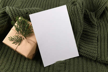 empty 5x7 card mockup kraft gift box with fir decoration on knitted green sweater, design element for wedding invitation, thank you or greeting card. Christmas winter card mock up backgroundの写真素材
