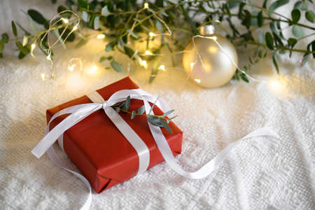Christmas red kraft gift box with white ribbon with eucaliptus leaves and light garland on a knitted beige cozy winter background. Cozy Christmas still life, birthday or new year, valentines day giftの写真素材