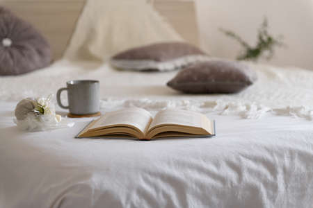 Cozy bedroom with bed covered of knited beige plaid, open a book, mug, cozy winter day at home. Christmas, relaxation and hygge background, coziness, comfort, interior and holidays conceptの写真素材