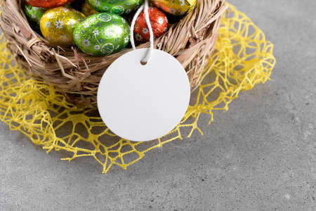 Round white Easter tag mockup with Happy Easter chocolate easter eggs in birds nest on gray stone background. Easter gift tag or sign for message greeting, close upの写真素材