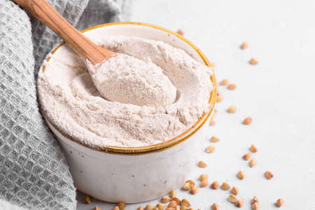 Buckwheat flour in a ceramic bowl with wooden spoon and raw green buckwheat grain on light gray concrete background, close up. Alternative flour, gluten free flour, healthy nutritionの写真素材