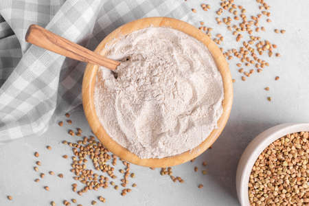 Buckwheat flour in a wooden bowl with wooden spoon and raw green buckwheat grain and napkin on light gray stone background, close up, top view. Alternative flour, gluten free flour, healthy nutritionの写真素材