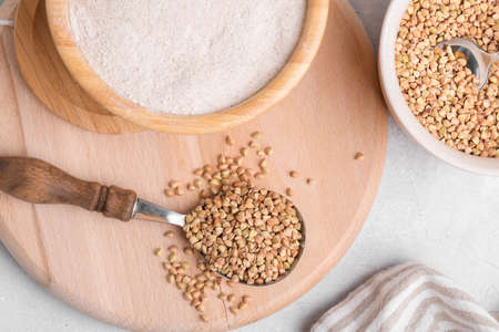 Raw green buckwheat grain in a spoon with buckwheat flour in a wooden bowl on light gray stone background, close up, top view, copy space. Alternative flour, gluten free flour, healthy nutritionの写真素材