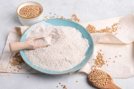 Buckwheat flour in a blu ceramic plate and in wooden scoop, raw green buckwheat grain on a bowl and napkin on light gray table background. Alternative flour, gluten free flour, healthy nutritionの写真素材