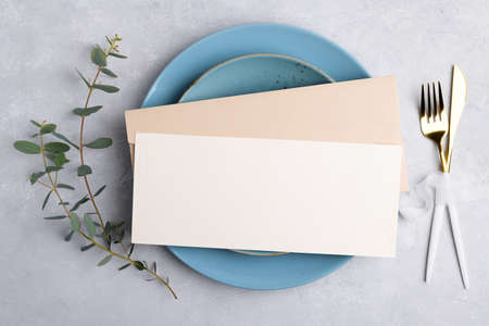 Invitation blank card mockup with festive or birthday table setting with golden cutlery, eucalyptus, blue ceramic plate on gray background. Restaurant menu concept. Flat lay, top viewの写真素材