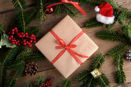 Christmas kraft gift box with red ribbon on rustic wooden background, Christmas present background with natural fir branches, cones, Cozy Christmas still life, new year, flat lay, top view