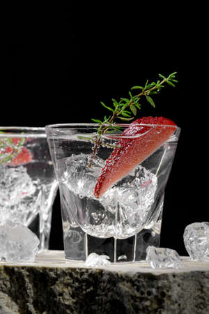 Vodka in two shot glass with ice, decorated with strawberry slice and thyme on travertine podium on dark stone background. Alcohol cocktail shot with strawberry and gin, fresh summer shots for partyの写真素材