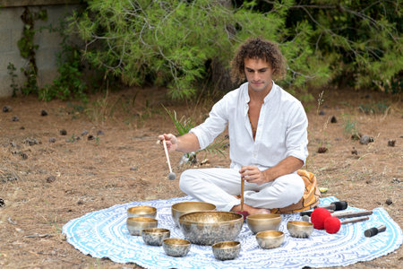 A man playing on a Tibetan singing bowls sitting outdoors for meditation. Buddhist healing practices. Sound therapy, alternative medicine. Clearing the space of negative energy. selective focusの写真素材