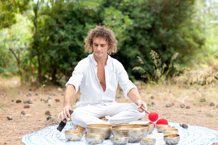 A man playing on a Tibetan singing bowls sitting outdoors for meditation. Buddhist healing practices. Sound therapy, alternative medicine. Clearing the space of negative energy. selective focusの写真素材